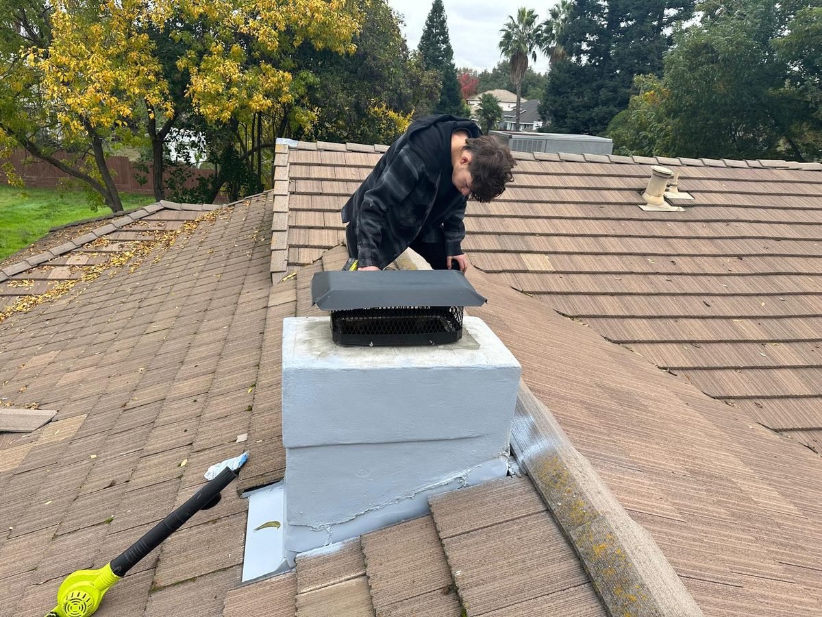 A technician fitting a black mesh chimney cap onto a painted chimney on a concrete tile roof