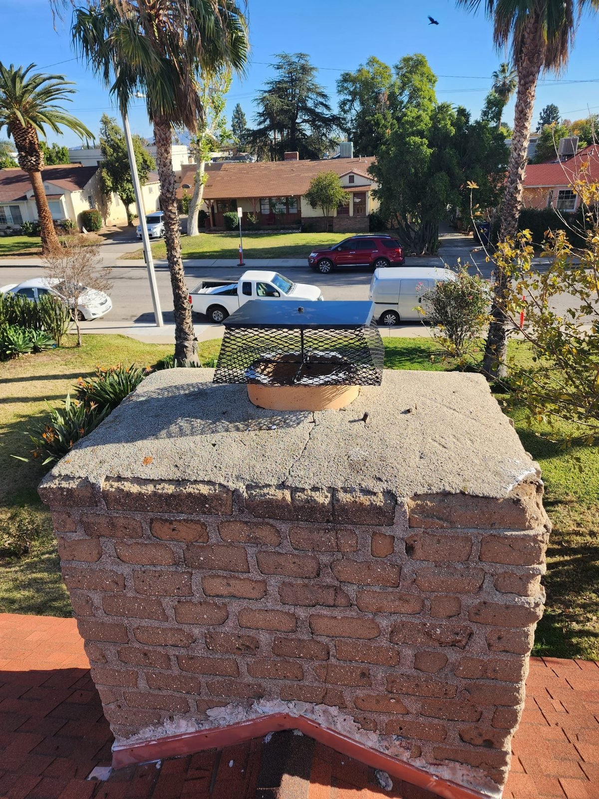 A cracked and weathered mortar chimney crown with a rain cap, photographed from the roof above a palm-lined street