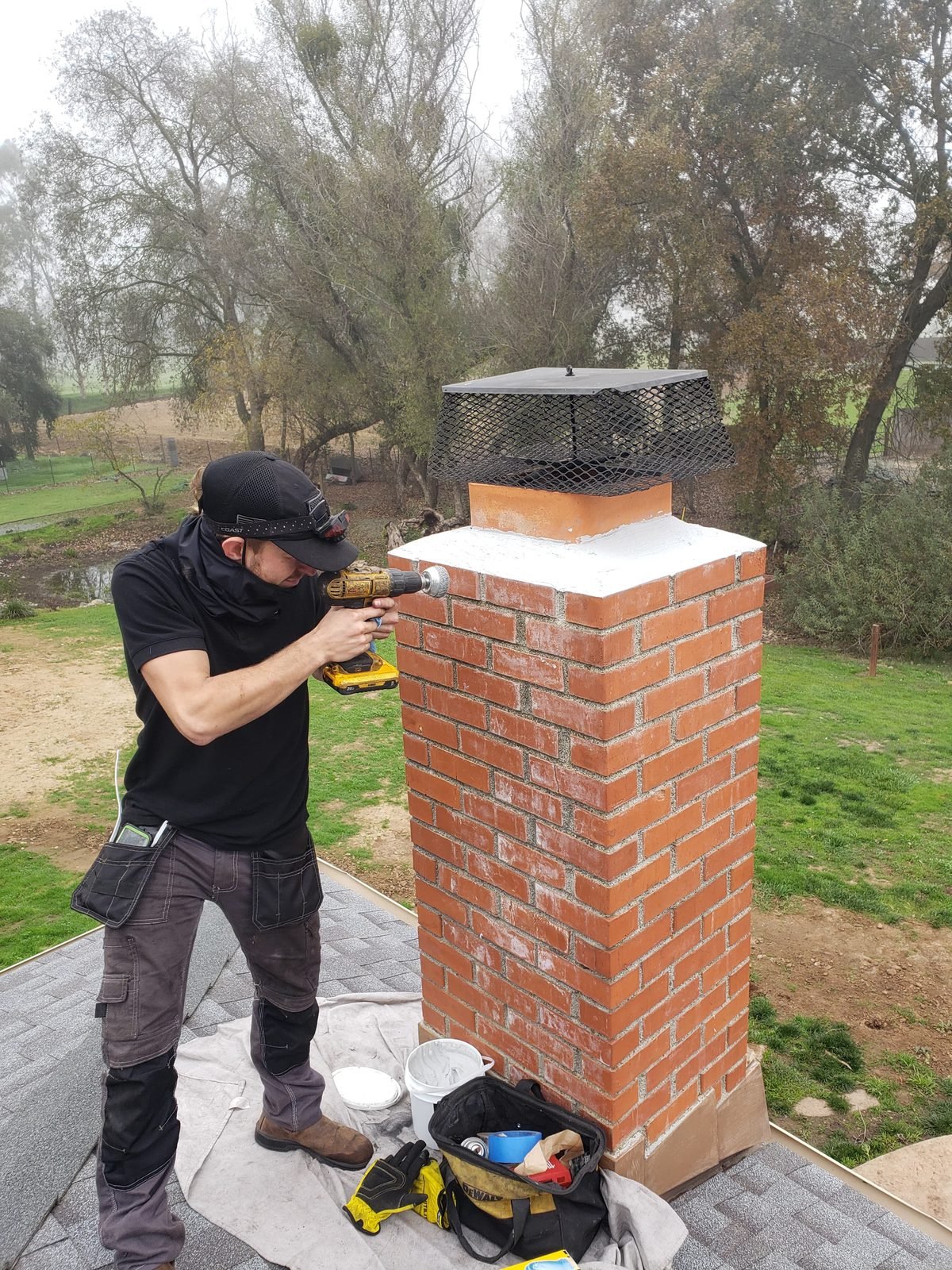 A technician on a roof drilling into a red brick chimney with a freshly finished white crown and a new cap fitted above