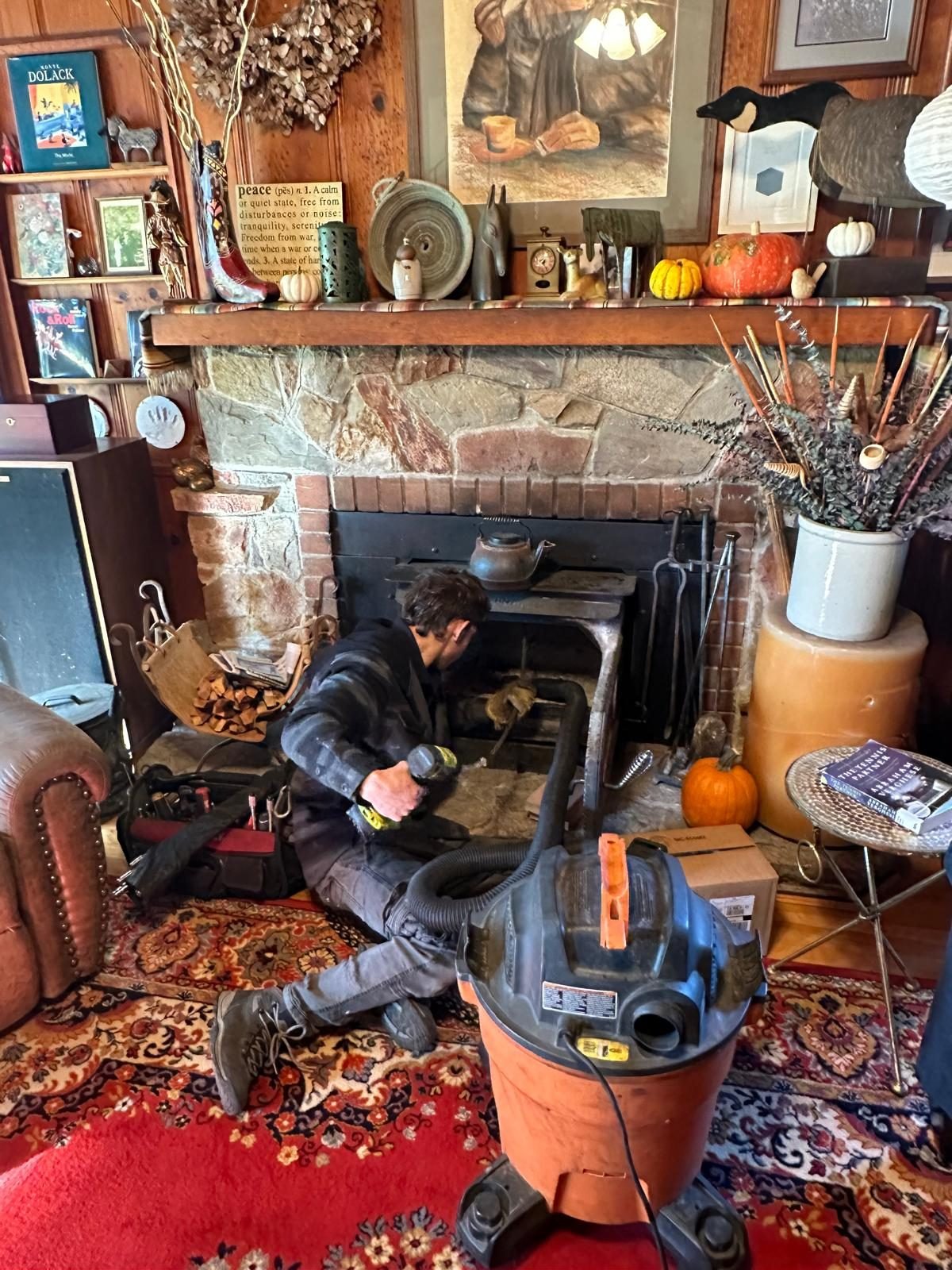 A technician kneeling at a stone fireplace working a brush into the flue, with a vacuum hose running to the hearth