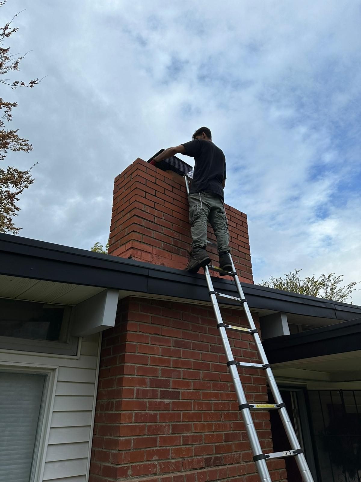 A technician on a telescoping ladder fitting a cap to the top of a tall red brick chimney