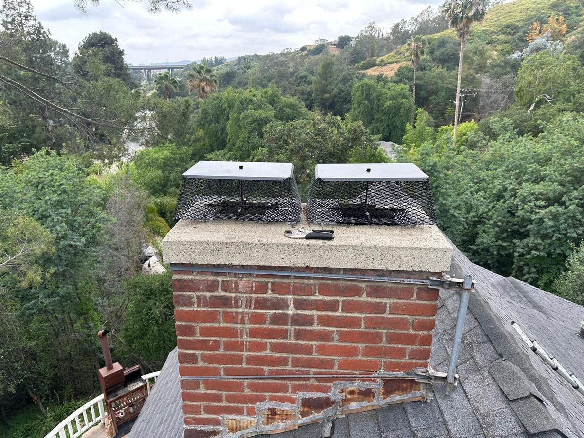 Two newly fitted mesh chimney caps side by side on a red brick chimney with a hillside behind